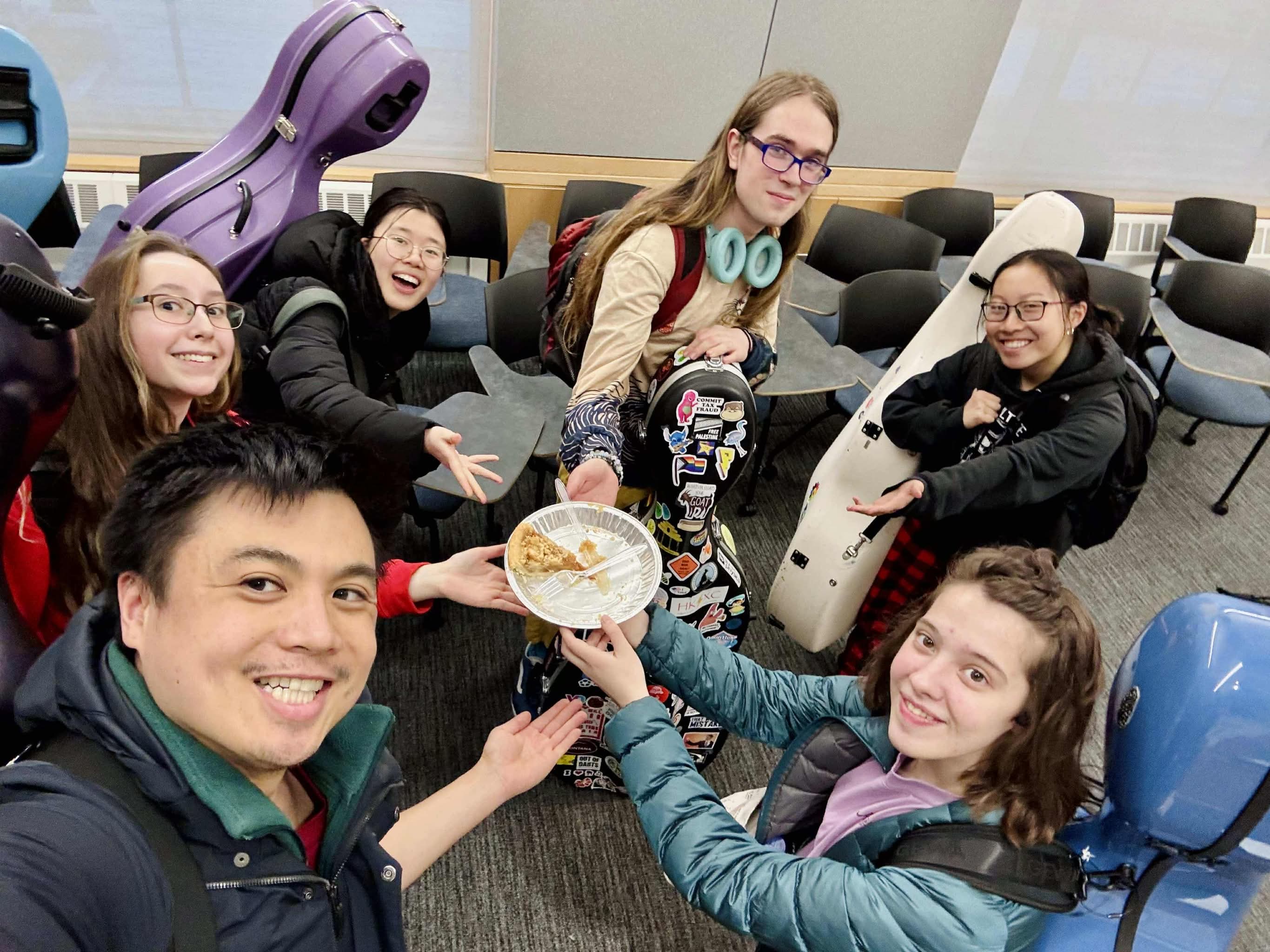 Celloworld members stand in a circle, holding aloft a tin containing the single remaining slice of pie after a Pi Day rehearsal.